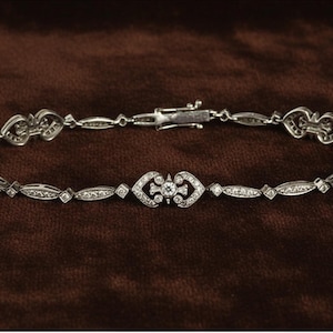 May include: A silver bracelet with a delicate design, featuring heart-shaped and oval links. The bracelet is adorned with small, sparkling gemstones. The clasp is visible, and the bracelet is set against a brown background.