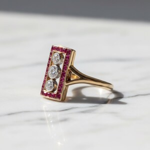 Pu&ograve; includere: Un anello in oro con testa rettangolare bordata da piccoli rubini rossi a taglio quadrato che circondano tre diamanti rotondi a taglio brillante allineati verticalmente in metallo bianco, con fascia in oro lucido e gambo sdoppiato.