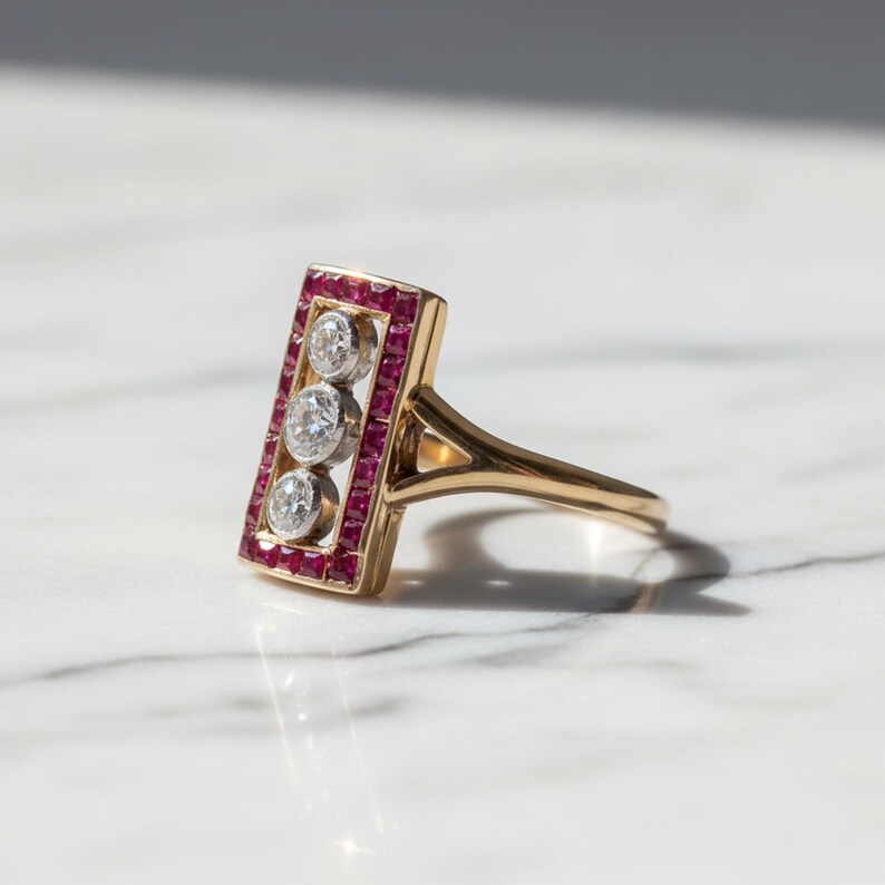 Pu&ograve; includere: Un anello in oro con testa rettangolare bordata da piccoli rubini rossi a taglio quadrato che circondano tre diamanti rotondi a taglio brillante allineati verticalmente in metallo bianco, con fascia in oro lucido e gambo sdoppiato.