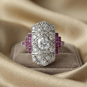 May include: A silver-toned ring featuring a central round diamond set within a geometric frame, surrounded by smaller round diamonds and accented by vertical rows of square-cut deep red rubies on both the left and right sides of the setting.