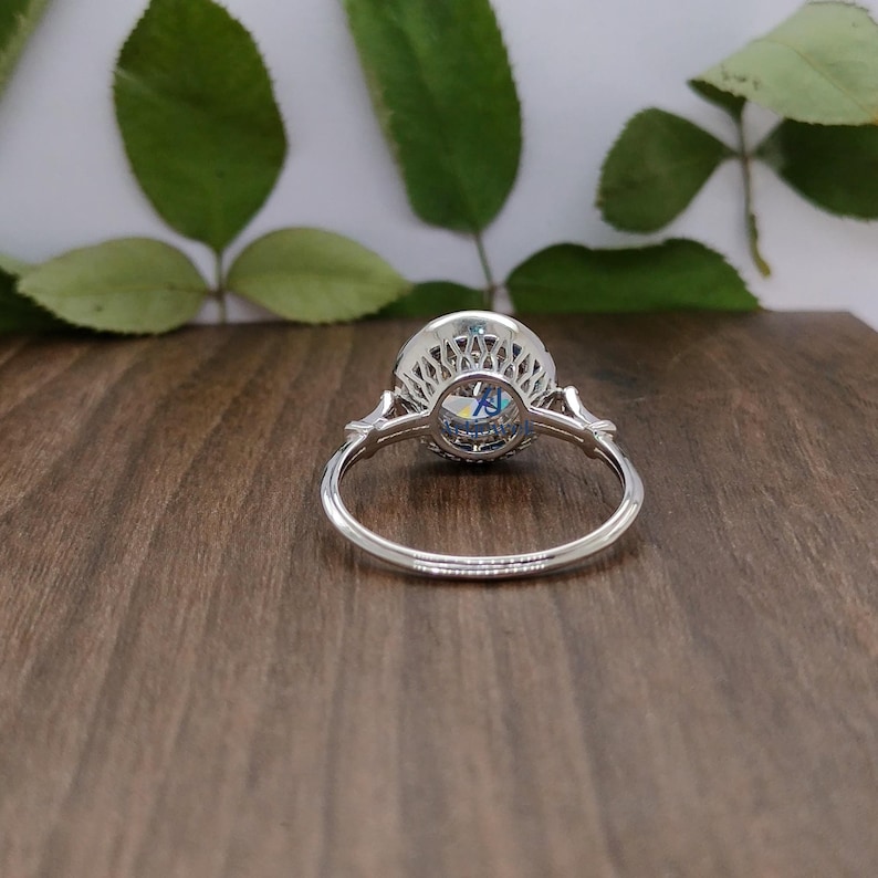 May include: A silver ring featuring a large, round, light blue gemstone at its centre, encircled by smaller stones. The ring showcases an intricate filigree design around the gemstone and on the shoulders of the band. It is set against a wooden surface.