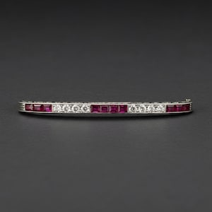 May include: A curved, silver-toned bar pin featuring alternating square-cut red gemstones and round, clear gemstones. The pin is set against a dark gray background.