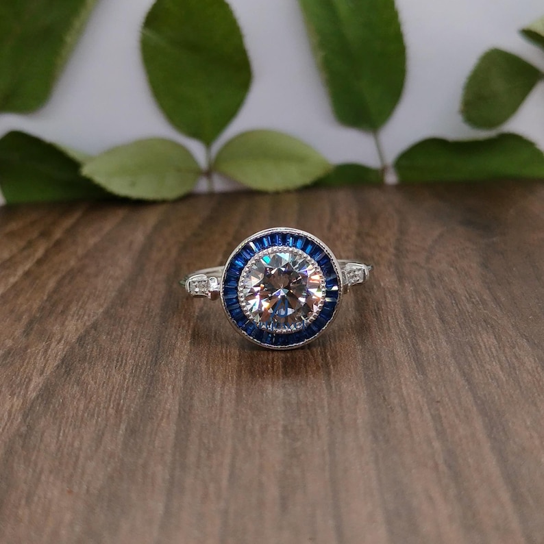 May include: A silver ring featuring a large, clear, round central stone encircled by a halo of small, blue rectangular stones. The ring is displayed on a dark wooden surface, with green leaves in the background.
