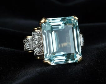 May include: An aquamarine-colored gemstone ring with a large, rectangular cut. The ring features gold accents on the corners and band, with intricate detailing and small, clear stones on the sides. The ring is set against a black background.