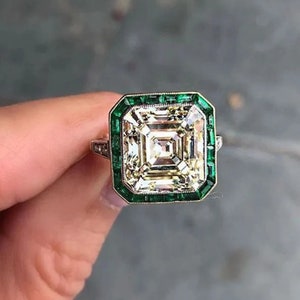 May include: A square-shaped engagement ring with a large, clear, faceted diamond center stone. The diamond is surrounded by a border of smaller, green emerald stones.
