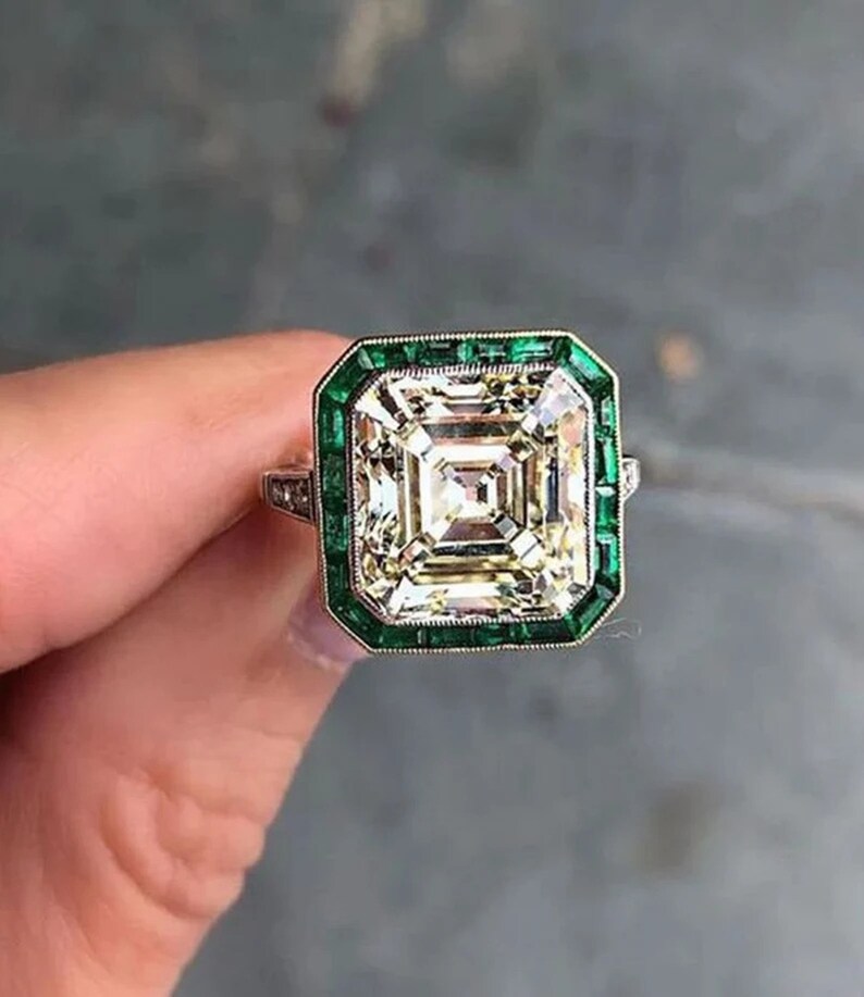May include: A square-shaped engagement ring with a large, clear, faceted diamond center stone. The diamond is surrounded by a border of smaller, green emerald stones.