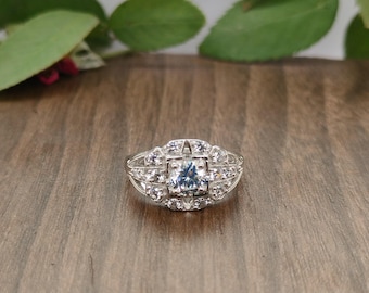May include: A silver ring with a square, light blue center stone, surrounded by smaller, clear stones. The ring has an ornate, vintage design, set against a wooden surface with green leaves in the background.