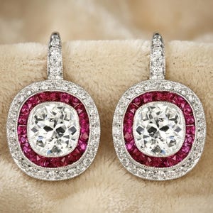 Puede incluir: Un par de pendientes elegantes con un diamante central de talla brillante, rodeado por un halo de gemas rojas vibrantes. El borde exterior está incrustado con piedras blancas más pequeñas y brillantes, todo engastado en metal plateado. Diseño clásico.