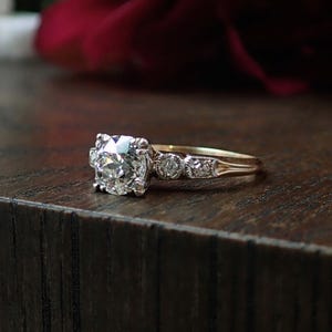Puede incluir: Un anillo de compromiso de oro con un gran diamante de talla redonda. El anillo tiene diamantes más pequeños en la banda y está sobre una superficie de madera oscura. Un diseño clásico.