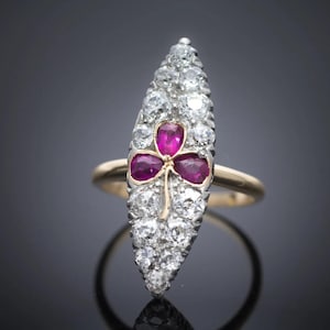 May include: A gold ring with a long, diamond-studded centerpiece. Three ruby gemstones form a clover shape at the center. The ring is set against a dark background, highlighting the sparkle of the diamonds and the rich color of the rubies.