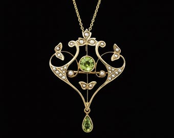 May include: A gold-toned pendant necklace with an ornate design. The pendant features a central round green gemstone, a teardrop-shaped green gemstone, and small white pearls. The necklace has a delicate chain and is set against a black background.