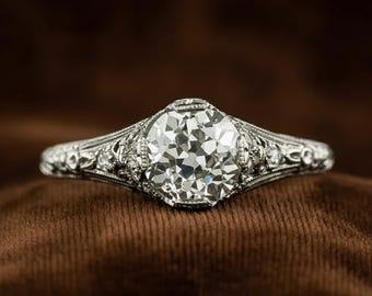 Könnte beinhalten: Ein Ring im Vintage-Stil mit einem großen, rund geschliffenen Mittelstein. Der Ring hat aufwendige Filigranarbeiten am Band und um die Fassung, mit kleineren Akzentsteinen. Der Ring liegt auf einer braunen Oberfläche.