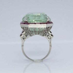 May include: A vintage ring featuring a large, light green gemstone surrounded by a halo of red rubies. The ring is set in platinum and has a delicate filigree design.