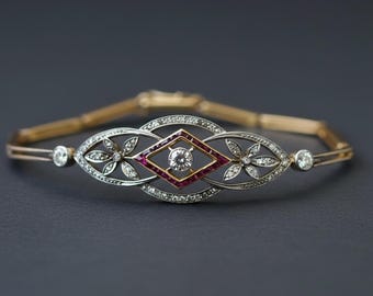 May include: An antique gold and silver bracelet with a central diamond and ruby design. The bracelet features a diamond-studded oval and floral design with a diamond in the center. The bracelet is approximately 7 inches long.