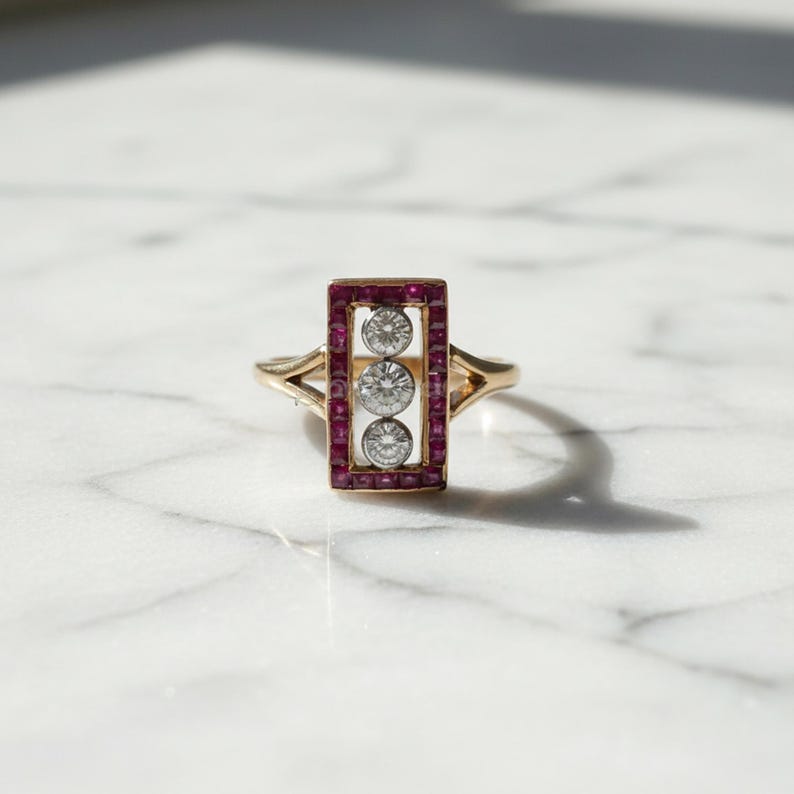 Pu&ograve; includere: Un anello in oro con testa rettangolare bordata da piccoli rubini rossi a taglio quadrato che circondano tre diamanti rotondi a taglio brillante allineati verticalmente su una barra centrale in metallo argentato con fascia a gambo diviso.