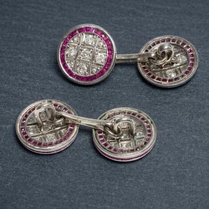 May include: A pair of silver cufflinks with a circular design. Each cufflink features a central square pattern of small, clear stones, surrounded by a ring of small, red stones. The cufflinks are connected by a silver bar.