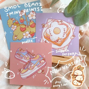 May include: Three small square prints featuring cute cartoon illustrations. The first print is on a blue background and features a strawberry plant with white flowers and red strawberries. The second print is on a purple background and features a plate with a slice of toast with a fried egg in the shape of a flower. The third print is on a pink background and features a pair of blue and yellow sneakers with small cartoon characters on them. The text "SMOL BEANZ: MINI PRINTS" is at the top of the image. The text "matte" is below the first print. The text "Smooth Surface" is below the second print. The text "NEW VERSION" is in a speech bubble with the text "free every 1 set" below it.