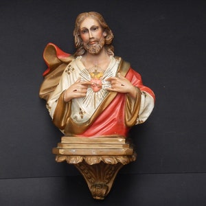 Puede incluir: Un busto de yeso de Jesucristo montado en la pared, con una túnica roja y dorada, las manos levantadas en un gesto de bendición, y un corazón con una corona de espinas en el pecho.