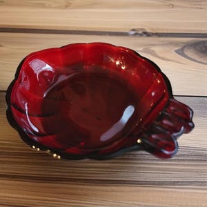 May include: A red glass ashtray with a scalloped edge and a gold rim. The ashtray has four indentations for cigarettes.
