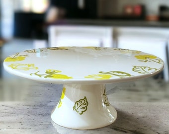 May include: White ceramic cake stand with a yellow and green floral design. The stand has a round base and a wide, flat top.