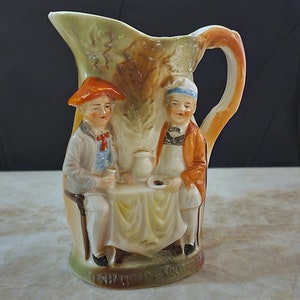 May include: A ceramic pitcher with a handle shaped like a tree trunk. Two figures are depicted on the pitcher, sitting at a table with a cup and saucer. The figures are wearing 18th-century clothing and the pitcher is decorated with a brown and green glaze.