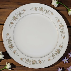 May include: A white porcelain dinner plate with a floral design. The plate features a delicate pattern of small, light brown and white flowers with green leaves around the rim. A thin silver band borders the inner edge of the plate.