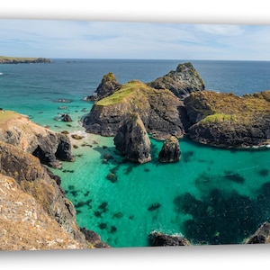 May include: A panoramic view of a rocky coastline with turquoise water. The water is clear and shallow, revealing the rocky bottom. The rocks are brown and grey, and there is some green vegetation growing on them.