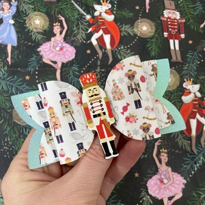 May include: A decorative hair bow with a central nutcracker figure in red, white, and gold, and patterned wings. The wings have a light blue glitter edge and a print of nutcrackers and other festive elements. The bow is held against a background with ballet dancers.
