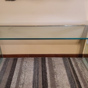 May include: A modern glass desk with a rectangular glass top and angled glass legs. The edges are trimmed with a gold-colored metal. The desk is set on a striped rug.