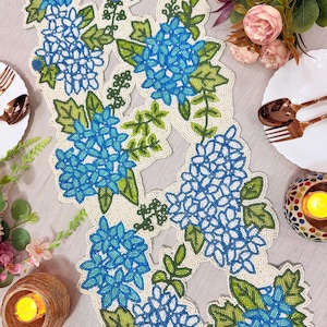 May include: A beaded table runner featuring a floral design with blue hydrangea blooms and green leaves. The runner is cream-colored with a beaded border and is set on a light gray surface. Decorative tableware and candles are in the background.