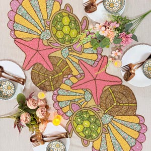 Pode incluir: Um caminho de mesa decorativo com tema marinho, apresentando estrelas do mar rosa, tartarugas verdes e desenhos de conchas amarelas e rosa. O caminho de mesa é rodeado por pratos brancos, talheres cor de cobre e arranjos florais.