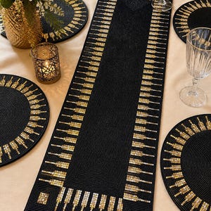 Op de afbeelding: Een rechthoekige zwarte tafelloper en vier bijpassende ronde placemats, allemaal voorzien van ingewikkeld zwart kraalwerk en decoratieve gouden en zilveren lovertjes die in een herhalend verticaal patroon langs de randen zijn geplaatst.