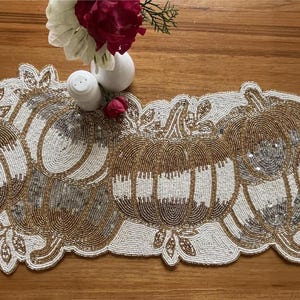 May include: A beaded table runner with a pumpkin design in gold and white. The runner has a scalloped edge and is decorated with leaves. Ideal for autumn decor.