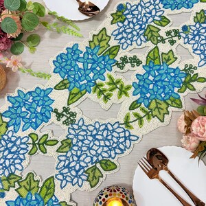 May include: A beaded table runner with a floral design featuring blue and white hydrangea blooms and green leaves. The runner is cream-colored with a beaded border. Two white plates with copper-colored forks and spoons are on either side.