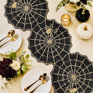 Könnte beinhalten: Schwarze, perlenbesetzte Spinnennetz-Tischsets mit goldenen Spinnenakzenten. Die Tischsets sind auf einem Tisch mit weißen Tellern, goldenem Besteck und Blumengestecken angeordnet. Halloween-Tischdekoration.