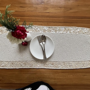 May include: A rectangular, off-white beaded table runner with a sequined border. A white plate with a fork and spoon sits on the runner. A small vase with red and white flowers is placed on the runner. The table is made of wood.