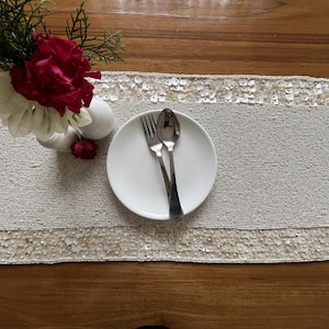 Op de afbeelding: Een ovale tafelloper met kralen en een witte paillettenrand. Een wit bord met vork en lepel ligt op de loper. Links staat een vaas met rode en witte bloemen. De loper ligt op een houten tafel.