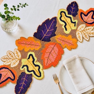 May include: A beaded table runner with a leaf design in autumnal colours. The runner features leaves in shades of orange, purple, yellow, and cream, with brown beaded detailing. A decorative piece for a dining table.