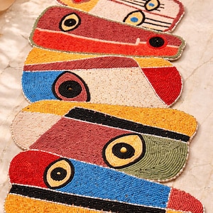 May include: A stack of colorful beaded placemats. Each mat features a unique design with geometric shapes and a large central eye-like motif. The color palette includes red, yellow, blue, and green, with black and white accents. The mats are oval-shaped.