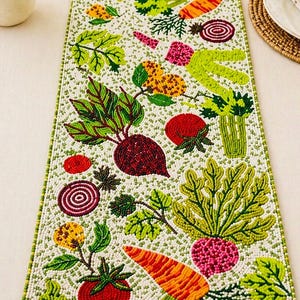 May include: A beaded table runner with a white background and a green border. The runner features colorful beaded vegetables, including carrots, beets, tomatoes, and leafy greens. The design is detailed and vibrant.