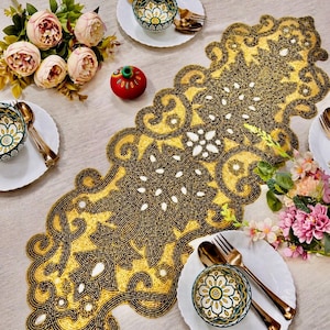 Op de afbeelding: Een goud-zwarte kralen tafelloper met een bloemmotief, gedekt met witte borden, gouden bestek en decoratieve kommen. De tafel is ook versierd met roze en crèmekleurige bloemen en een rood ornament.