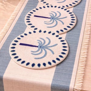 Op de afbeelding: Drie ronde placemats met een palmboomontwerp en een blauwe rand liggen op een blauw-wit gestreepte tafelloper. De placemats hebben een crèmekleurige achtergrond en blauwe accenten. De tafelloper heeft een gefranjerde rand.