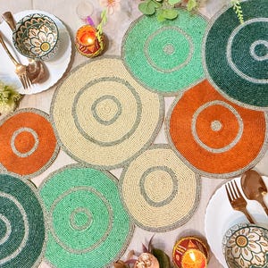 May include: A collection of beaded placemats in various colors, including green, orange, and cream. The placemats are circular with concentric circle designs. The image also includes plates, bowls, and cutlery, suggesting a dining setting.