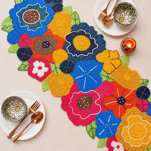 May include: A beaded floral table runner in vibrant colors, including blue, yellow, pink, and orange. The runner features a variety of flower shapes and green leaves. Two small bowls, a fork, and a spoon are on white plates.