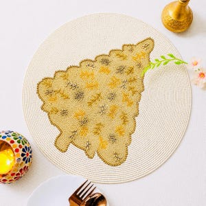 May include: Cream-colored beaded placemat with a gold and beige leaf design. The placemat has a scalloped edge and is surrounded by a woven border. A gold candle holder and a small floral arrangement are also visible.