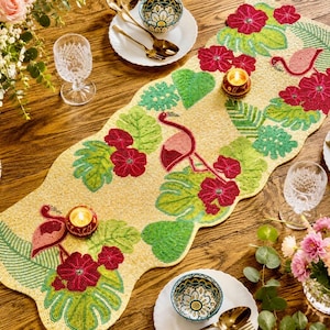 May include: Beaded table runner with a tropical design featuring flamingos, red flowers, and green leaves. The runner is yellow and has a scalloped edge. Candles and tableware are also visible.
