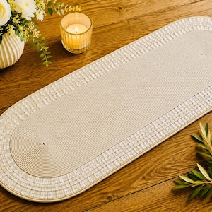 Peut inclure: Un chemin de table de forme ovale fabriqué à partir de corde beige enroulée, présentant une section centrale plate et une bordure décorative composée de petits carreaux blancs carrés disposés en grille sur tout le pourtour du chemin.