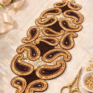 May include: A decorative table runner with a paisley design. The runner is brown with gold and bronze accents. The paisley shapes are outlined in gold beads. The background is a light beige with gold sequins.