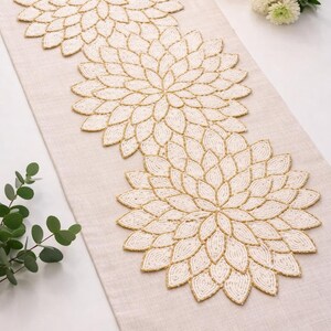 May include: Beige table runner with three large, beaded floral designs in cream and gold. The runner is on a light-colored surface, with a candle and flowers in the background. The floral designs are approximately 12 inches in diameter.