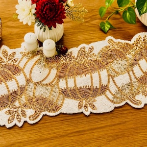 Op de afbeelding: Een kralen tafelloper met pompoenen in goud en wit, met een geschulpte rand. De loper is versierd met pompoenvormen en bladeren. Een kaars, bloemen en zout- en peperstrooiers staan op tafel.